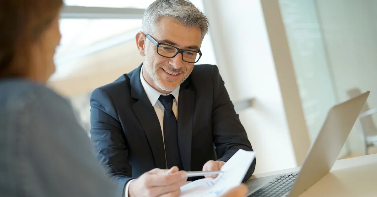  A middle-aged businessman in a suit and glasses discusses a document with a colleague across the table in a bright office.