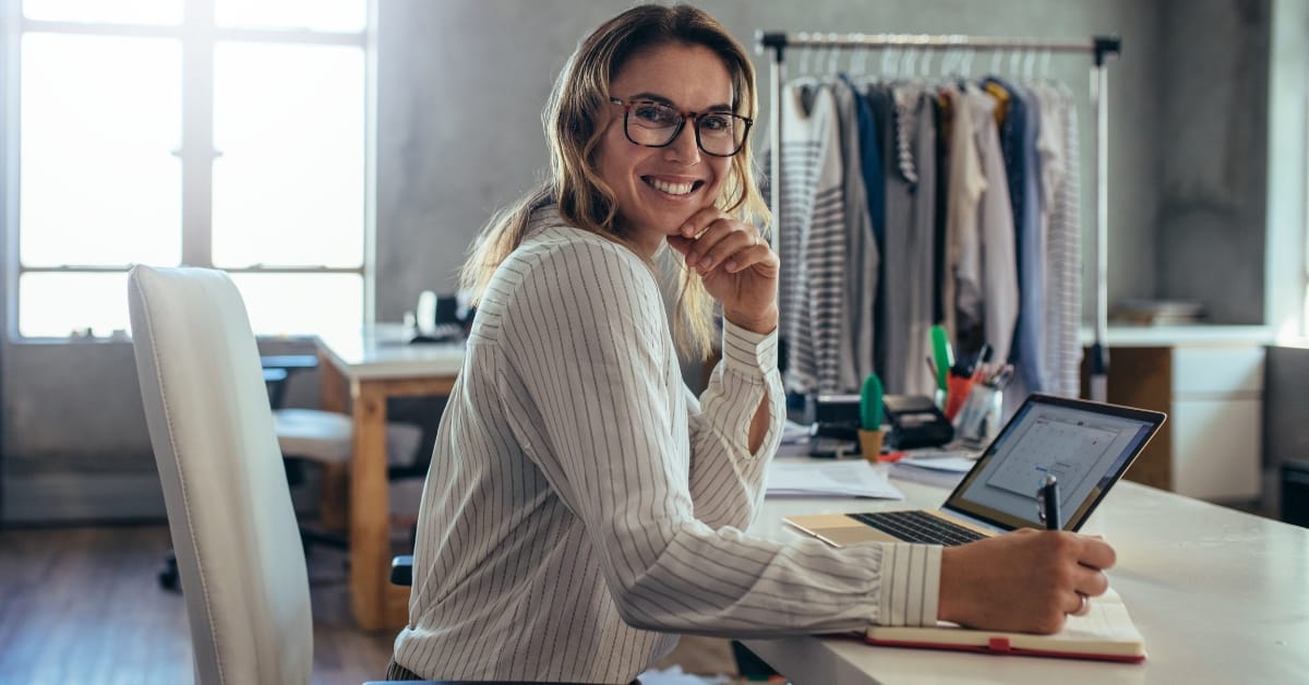 A smiling woman in a striped shirt works on a laptop in a creative workspace, surrounded by clothing racks and papers.