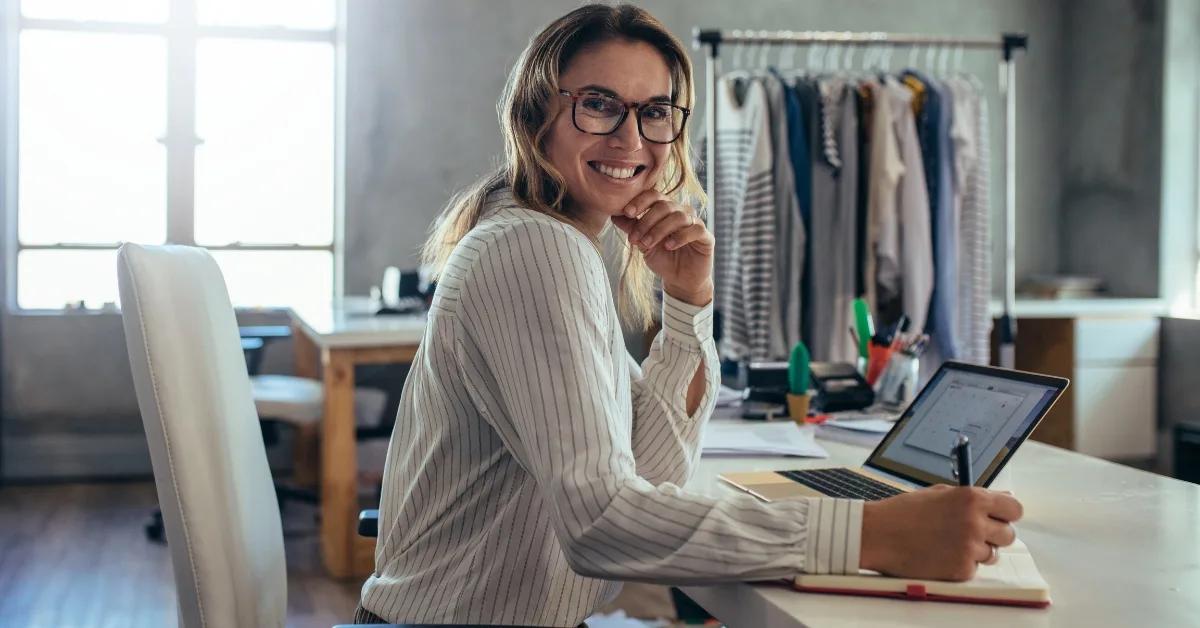 A smiling woman in a striped shirt works on a laptop in a creative workspace, surrounded by clothing racks and papers.