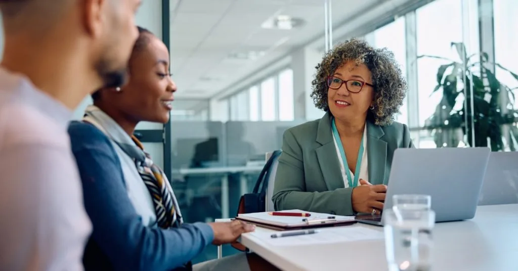 A businesswoman with curly hair and glasses smiles while meeting with a couple in a modern office setting, with a laptop and notepad on the table.