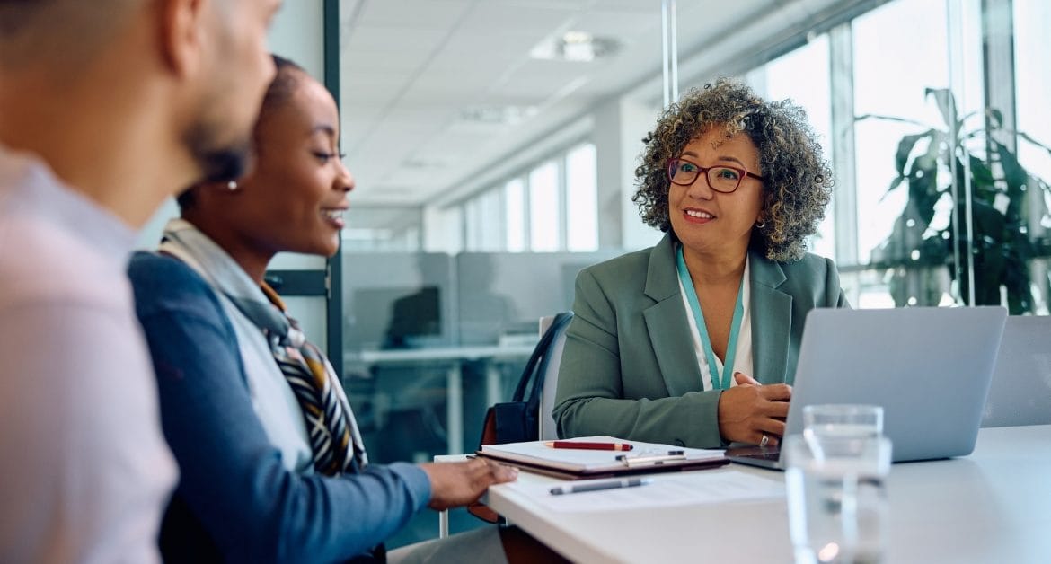 A businesswoman with curly hair and glasses smiles while meeting with a couple in a modern office setting, with a laptop and notepad on the table.