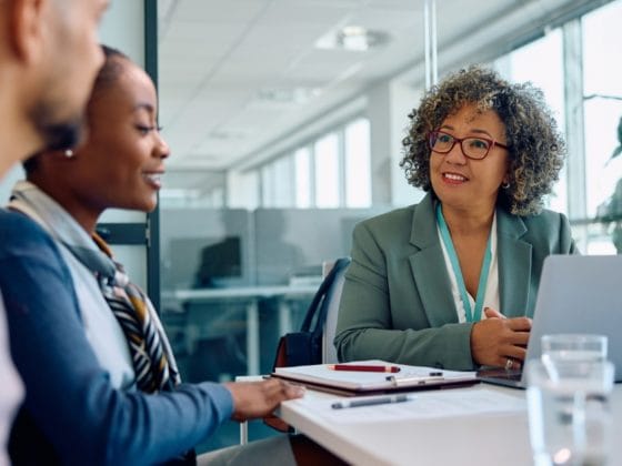 A businesswoman with curly hair and glasses smiles while meeting with a couple in a modern office setting, with a laptop and notepad on the table.