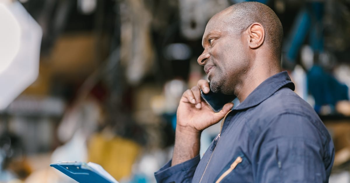 car repair delays due: A garage worker speaking on a mobile phone while holding paperwork in a busy workshop, suggesting delays or scheduling issues.