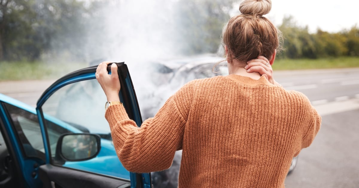 vehicle insurance write-offs: A driver standing beside a damaged car at the roadside, looking at smoke coming from the vehicle after a collision.