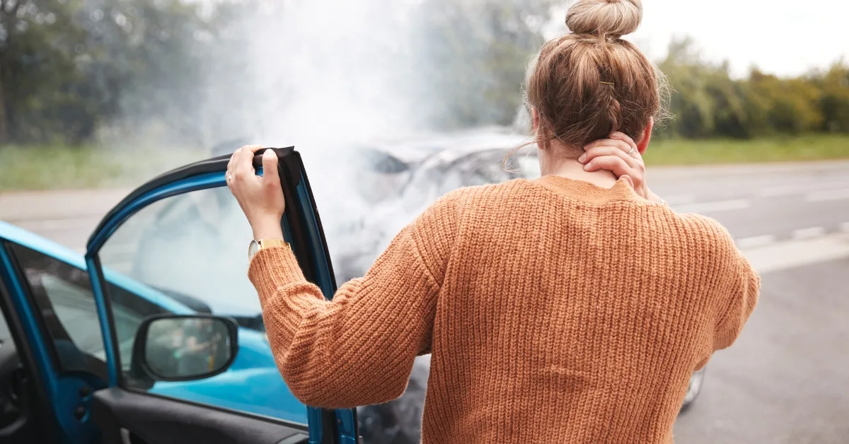 vehicle insurance write-offs: A driver standing beside a damaged car at the roadside, looking at smoke coming from the vehicle after a collision.
