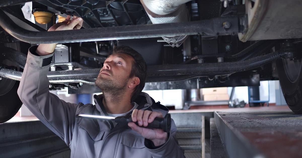 Car repair payment options: A male mechanic in a grey work jacket inspecting the undercarriage of a raised vehicle using a flashlight in a professional garage.