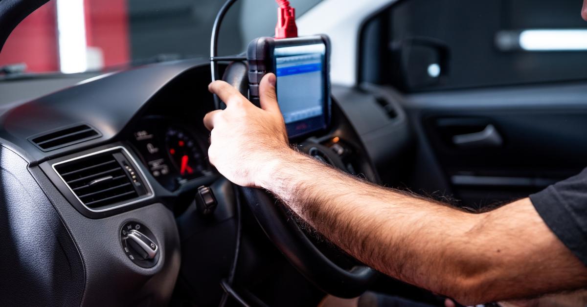 Professional car repair costs: A mechanic's arm reaching into a car interior to operate an OBD diagnostic scanner mounted on the steering wheel, with warning lights visible on the dashboard.