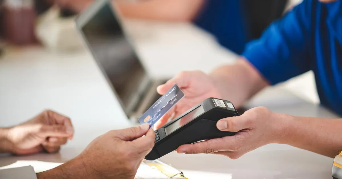 Car Repair Payment Options: A customer taps a credit card on a card payment terminal held by a garage staff member, with a laptop visible in the background.