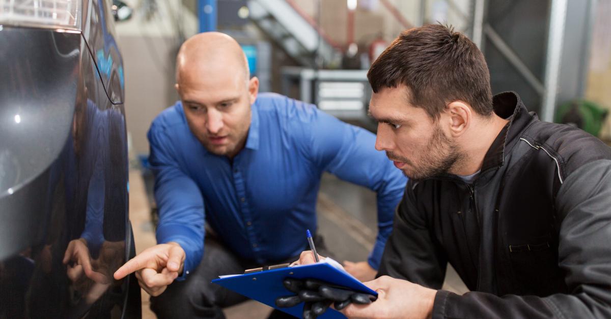 Car repair quality control: Two men crouch down to inspect the bodywork of a dark-coloured car in a garage workshop, one pointing at the vehicle while the other takes notes on a blue clipboard.