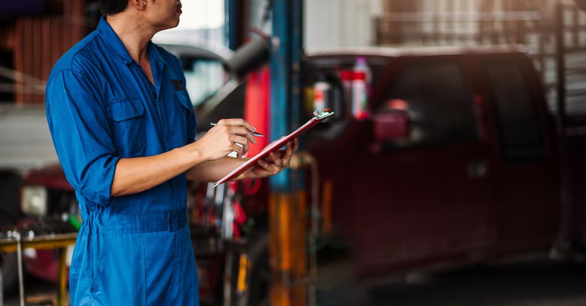 Cost of rework in garages: A mechanic in blue overalls stands in a garage holding a red clipboard and pen, with a vehicle on a lift in the background.