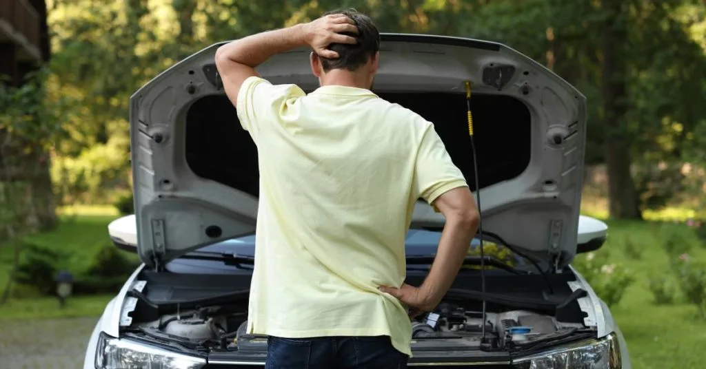 DIY car repairs: A man in a yellow t-shirt stands with his back to the camera, scratching his head in confusion whilst looking into the open bonnet of a car outdoors.