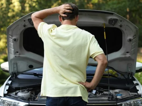 DIY car repairs: A man in a yellow t-shirt stands with his back to the camera, scratching his head in confusion whilst looking into the open bonnet of a car outdoors.