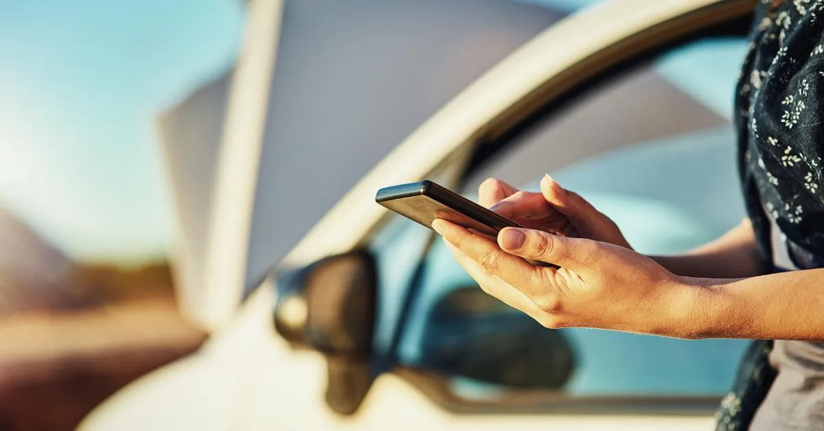 Dangers of DIY car repairs: A woman stands beside a car with its bonnet open, looking at her mobile phone in both hands on a sunny day.