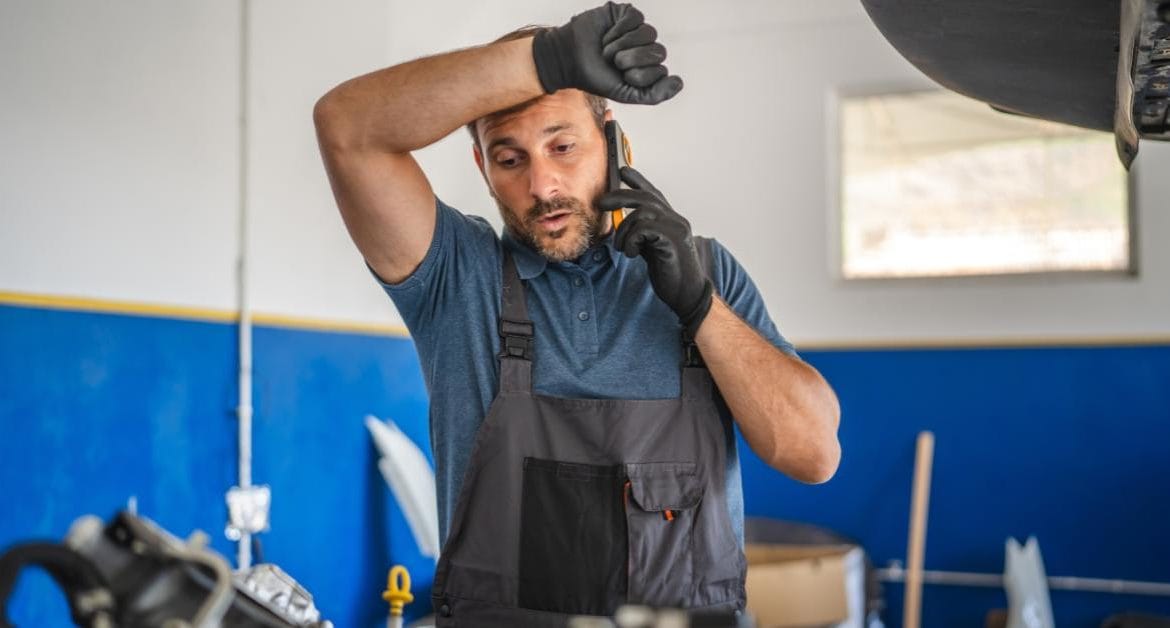 Workshop Quality Control: A stressed-looking mechanic in grey overalls and black gloves talks on a mobile phone whilst resting his hand on his forehead, standing in a garage with car parts visible around him.