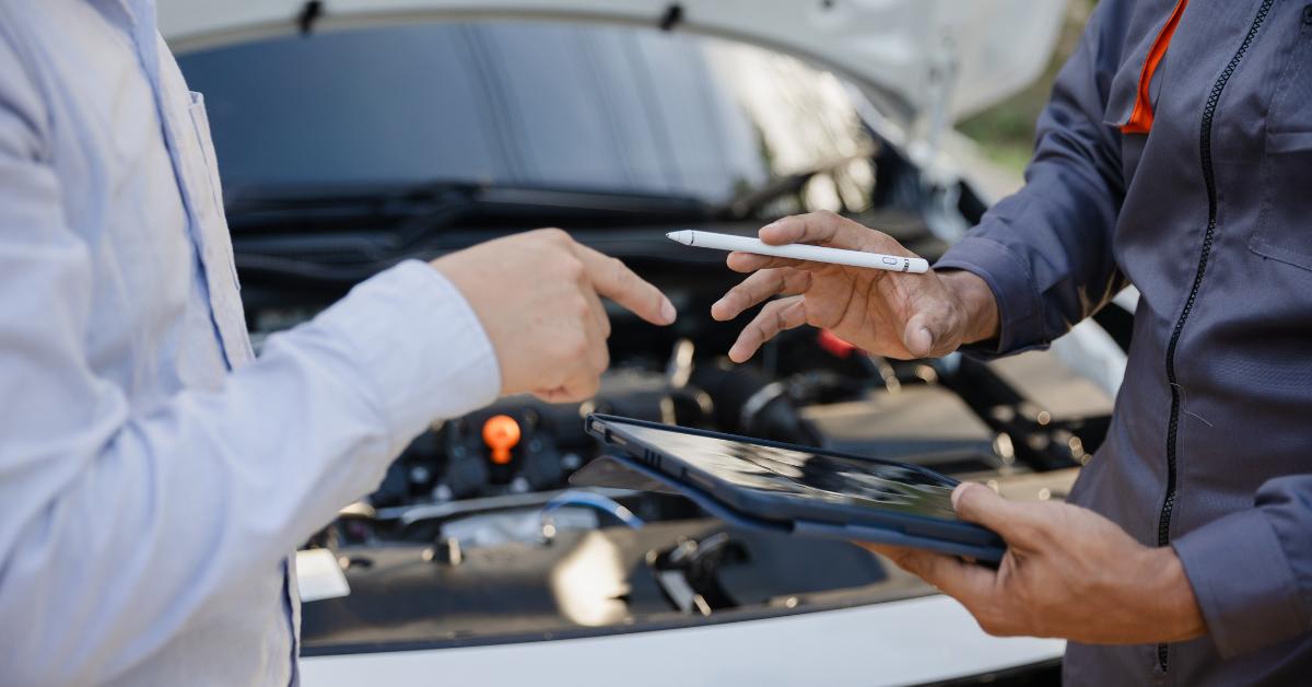 Car repair finance paying mechanic: A mechanic in a grey uniform holding a tablet and stylus whilst discussing repair details with a customer in front of an open car bonnet.