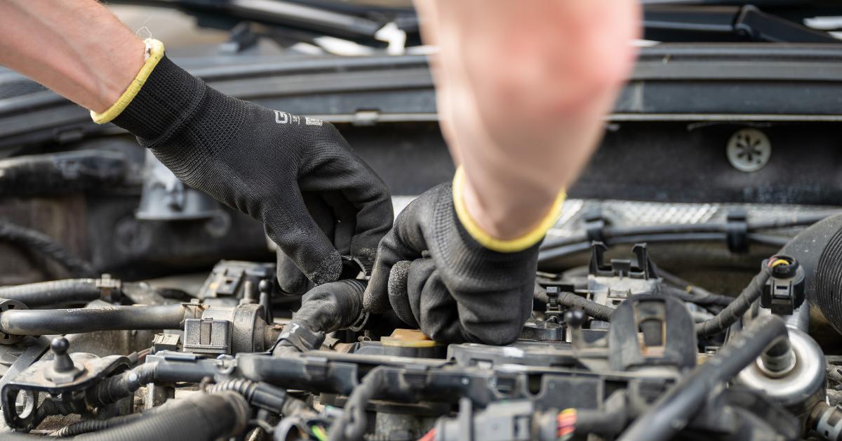 Car repairs after finance: A close-up of a mechanic's gloved hands working on a car engine, manipulating wiring and components within an open engine bay.